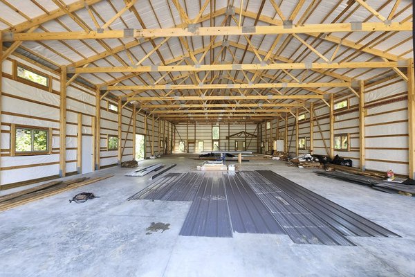 Interior view of a post-frame construction building by Built-Mor Buildings, featuring a spacious, open layout with exposed wooden trusses supporting the roof. The structure has insulated walls and large, evenly spaced windows allowing natural light. Build