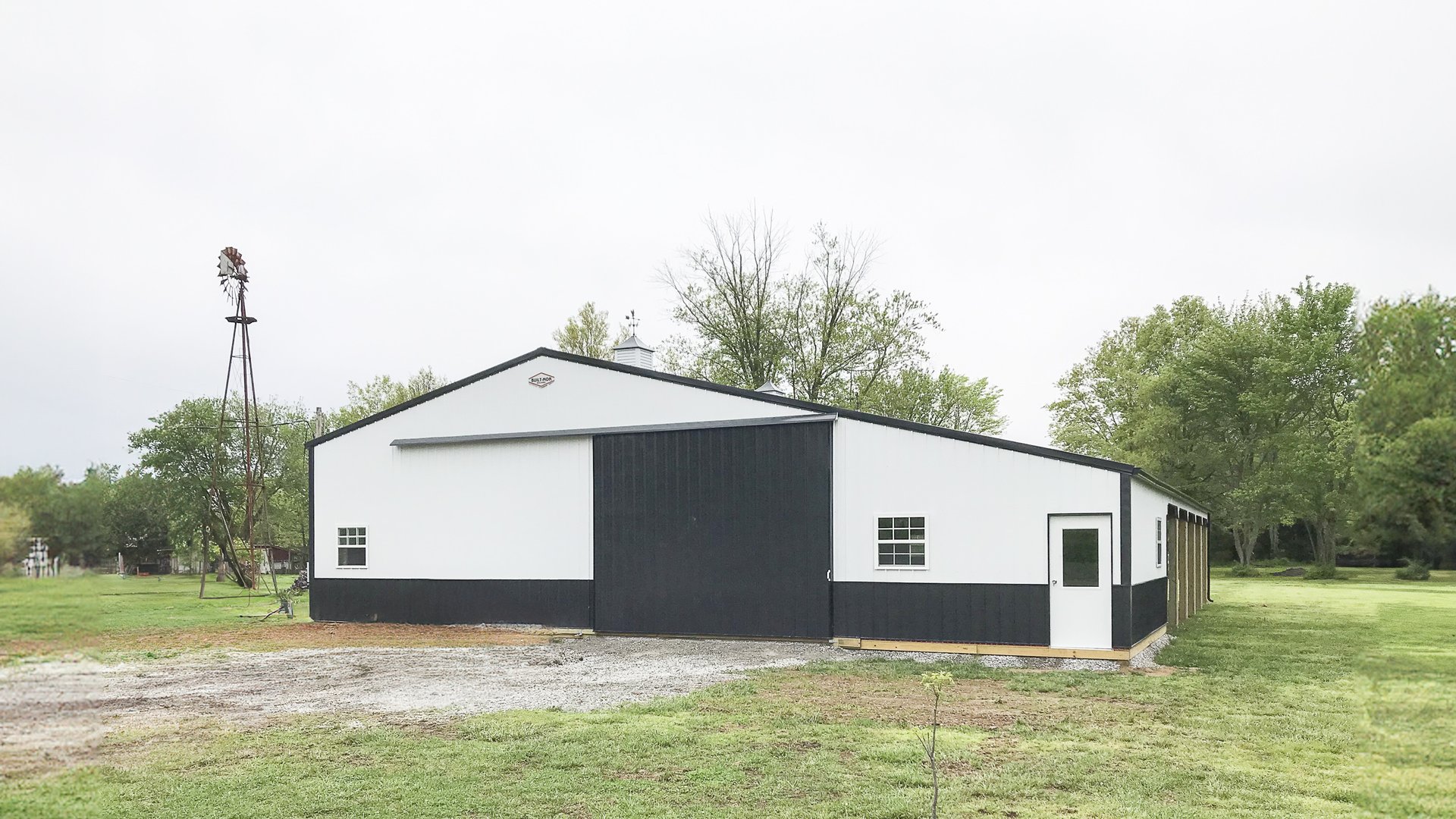 A post-frame construction building by Built-Mor Buildings is shown in a rural setting. The structure has a classic barn-like design with a gable roof and large sliding doors on the front facade. It features a two-tone color scheme with white upper walls a