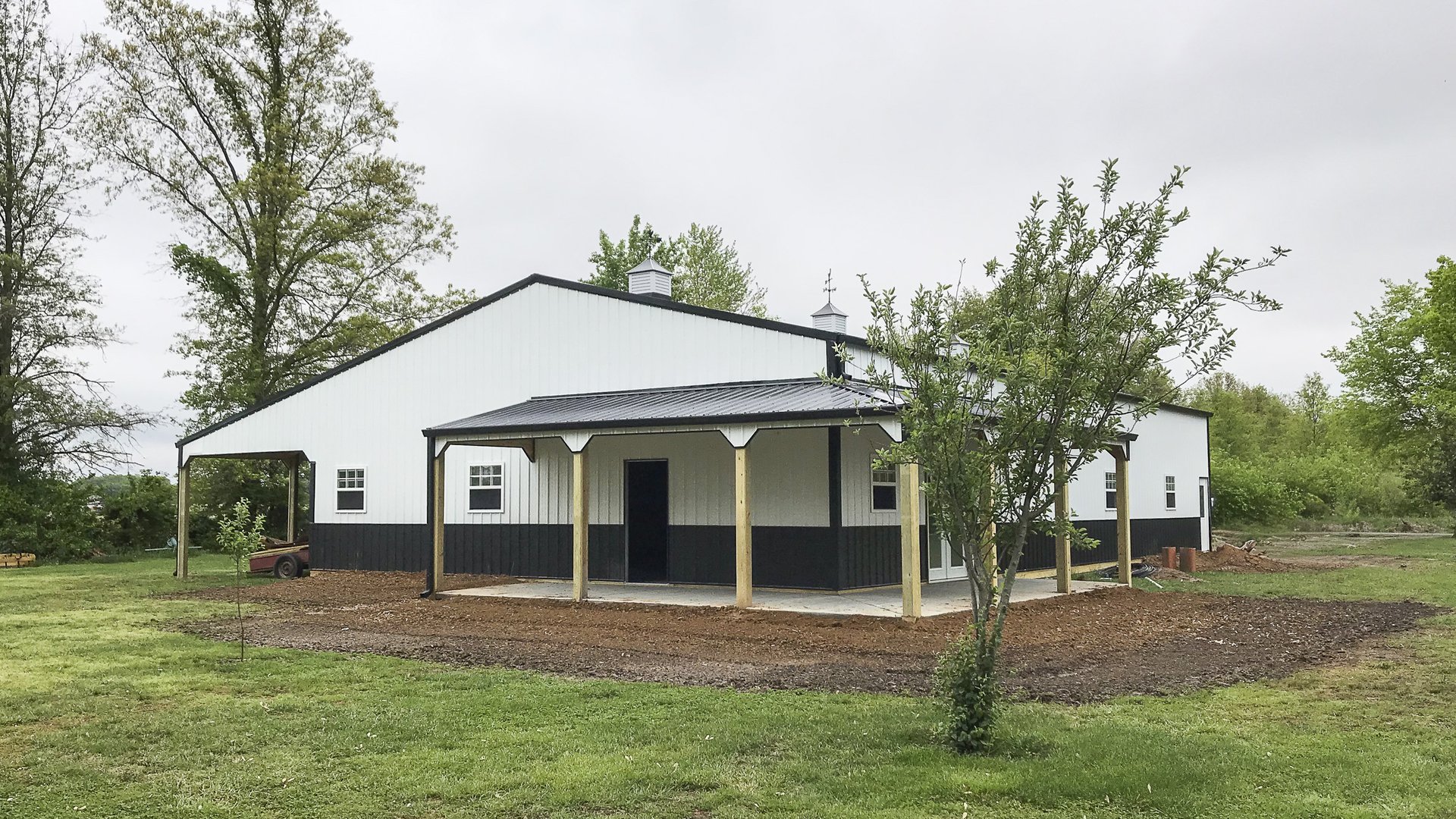 The image depicts a modern post-frame building set in a grassy area. The structure features a gable roof with two cupolas and a covered porch area supported by wooden posts. The building has a clean, two-tone exterior, with white upper walls and black low