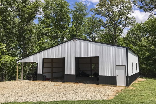 This image shows a post-frame building from Built-Mor Buildings, set against a wooded background. The structure features a white exterior with dark lower trim and includes three large open bays and a single-entry door, suggesting use as a garage or storag