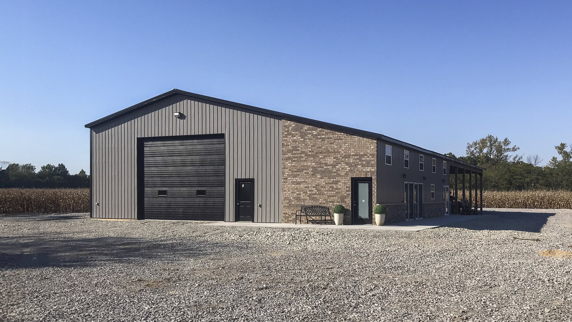 A modern post-frame building with a wide rectangular structure is depicted, featuring a high gabled roof. The exterior includes a section of dark grey metal siding complemented by brick detailing on the facade. Prominent elements include a large overhead