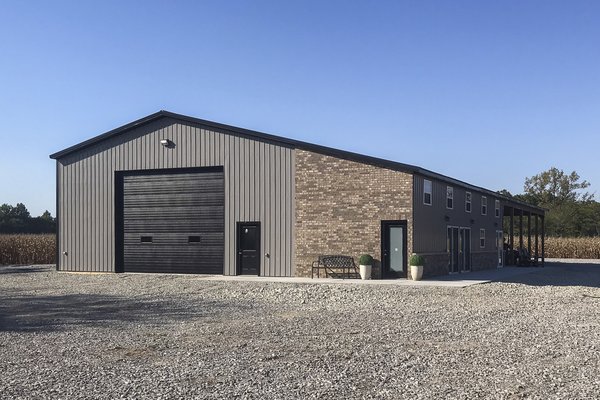 A modern post-frame building with a wide rectangular structure is depicted, featuring a high gabled roof. The exterior includes a section of dark grey metal siding complemented by brick detailing on the facade. Prominent elements include a large overhead