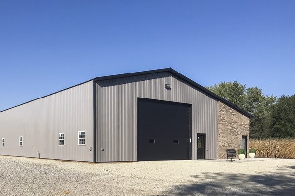 The image depicts a large post-frame building from Built-Mor Buildings, featuring a steel exterior and a gable roof. The front facade has a large black roll-up door and several white-framed windows. An entry door is adjacent to the roll-up door. The struc