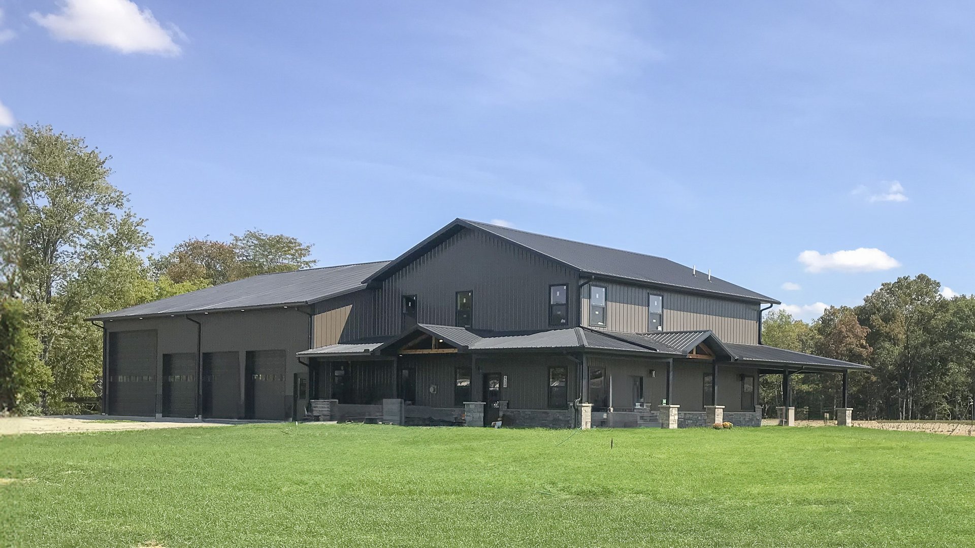 A large post-frame construction building with a dual purpose: part residential and part garage. The structure features a dark metal exterior with multiple sloped roofs. The residential section has several windows and an extended porch with stone pillars.