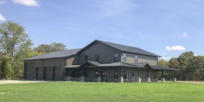 A large post-frame construction building with a dual purpose: part residential and part garage. The structure features a dark metal exterior with multiple sloped roofs. The residential section has several windows and an extended porch with stone pillars.