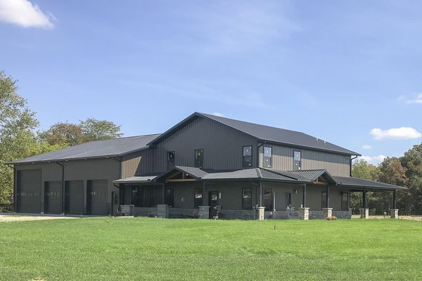 A large post-frame construction building with a dual purpose: part residential and part garage. The structure features a dark metal exterior with multiple sloped roofs. The residential section has several windows and an extended porch with stone pillars.