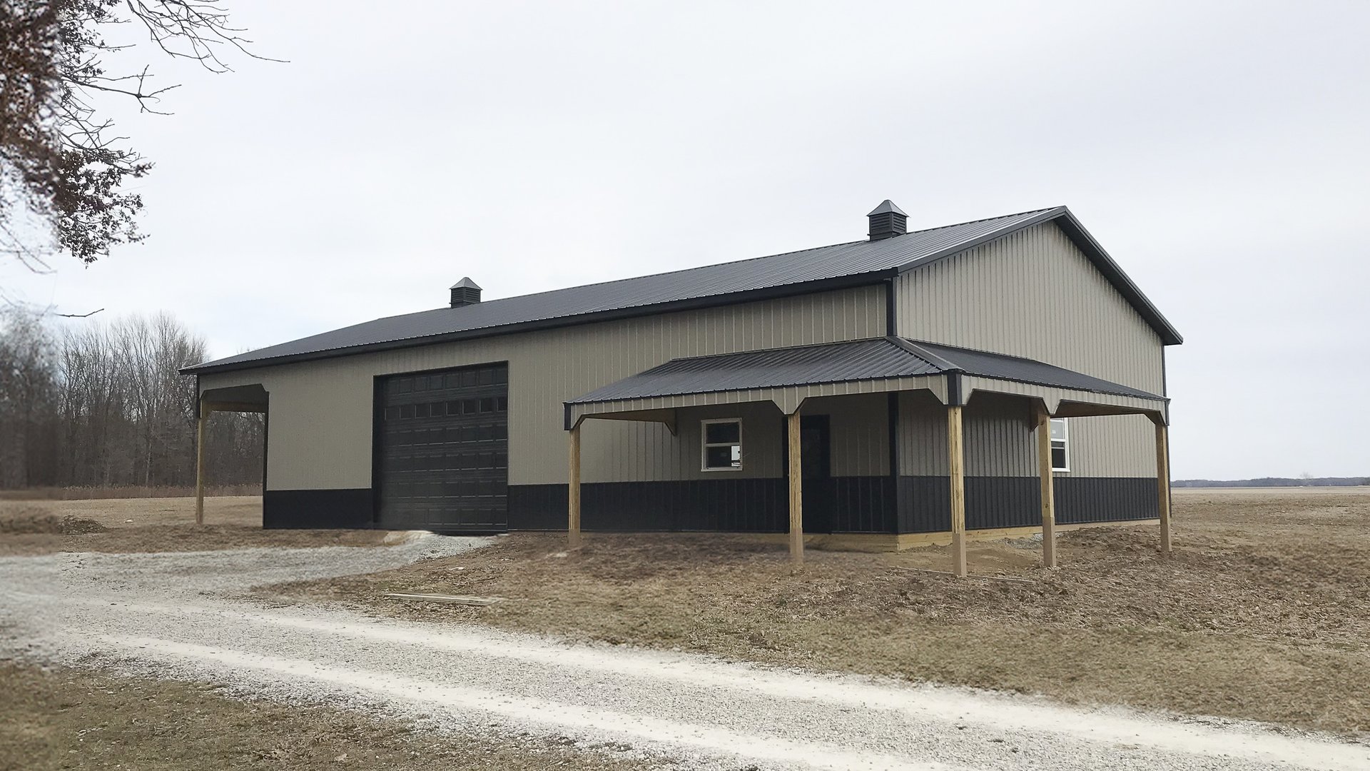 A post-frame construction building by Built-Mor Buildings is shown in a rural setting. The structure features a gable roof with two cupolas for ventilation and a combination of light and dark siding for contrast. A large overhead garage door suggests use