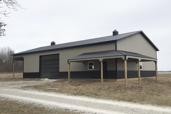 A post-frame construction building by Built-Mor Buildings is shown in a rural setting. The structure features a gable roof with two cupolas for ventilation and a combination of light and dark siding for contrast. A large overhead garage door suggests use