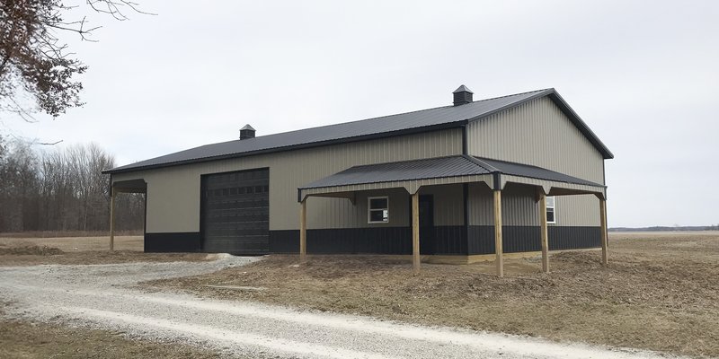 A post-frame construction building by Built-Mor Buildings is shown in a rural setting. The structure features a gable roof with two cupolas for ventilation and a combination of light and dark siding for contrast. A large overhead garage door suggests use