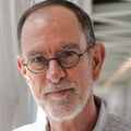 Michael Riscoe, Ph.D. has short dark gray hair, eye glasses, is tall, and wearing a light blue button-up shirt, standing on an indoor connecting bridge at OHSU.