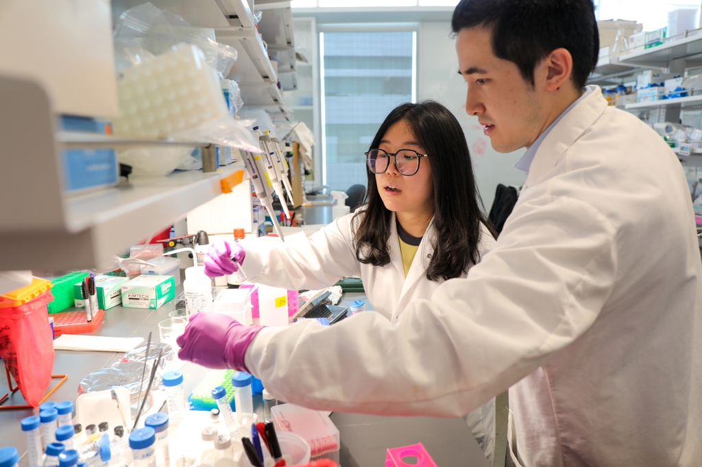 Image is of Li Xiang, who has long black hair, eye glasses, wearing a white coat, as she holds compounds at a research bench with Michael Henderson, who has dark hair, and a white coat. Li Xiang, Ph.D., postdoctoral scholar at the OHSU Knight Cancer Institute, and Michael Henderson, B.A., biomedical engineering Ph.D. student, are part of a team that developed a new nanoparticle that makes cancer treatment safer and more effective. (OHSU/Christine Torres Hicks)