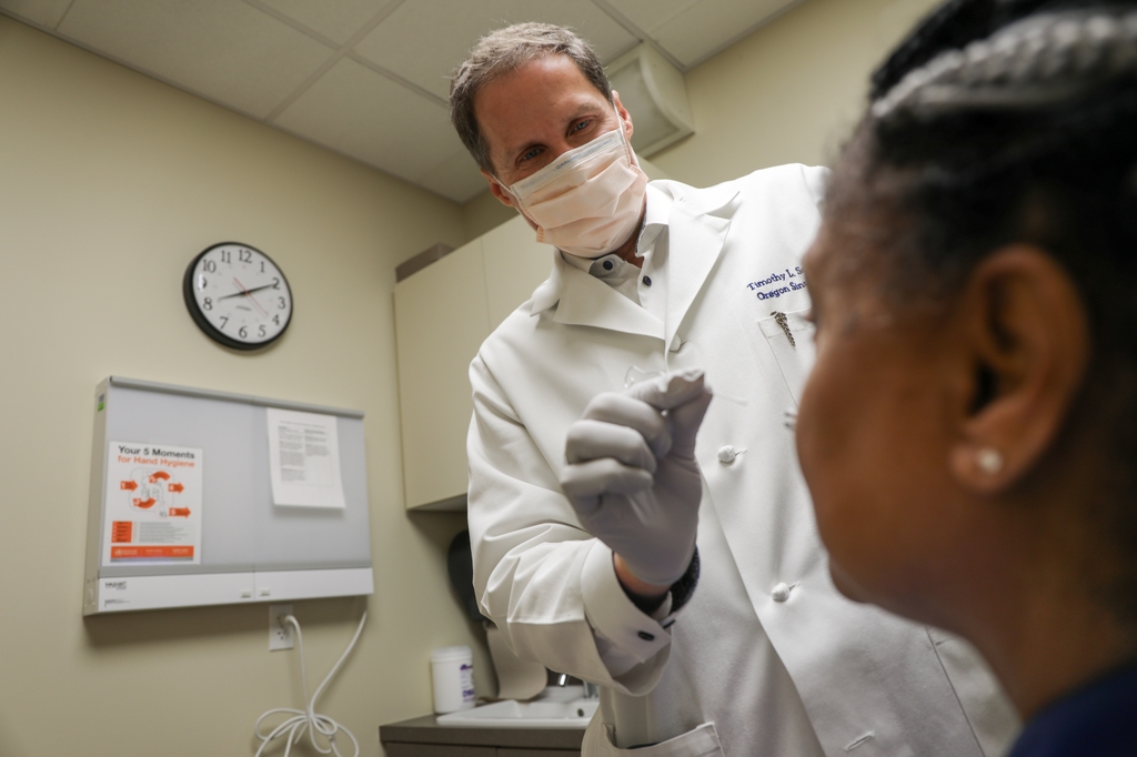 Photos shows Timothy Smith with gray hair and a white coat, face mask and gloves, about to put a swab up a patient's nose in an exam rooom. Timothy Smith, M.D., M.P.H., professor and interim chair of otolaryngology, is the lead principal investigator on a new NIH-funded grant to study treatments for chronic sinusitis. (OHSU/Christine Torres Hicks)