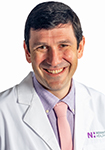 Dr. John Rommel is wearing a white lab coat over a purple shirt and pink tie and is smiling at the camera