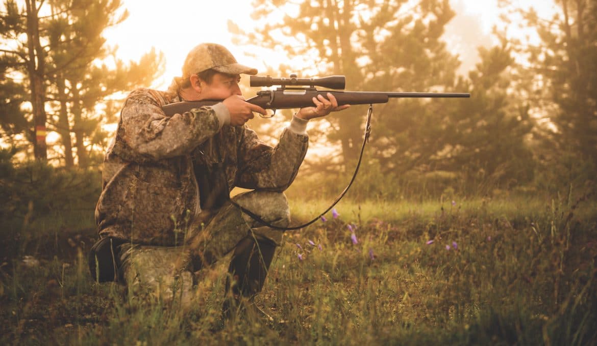 Portrait of a hunter while crouching and aiming with rifle at his prey.