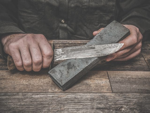 Man's hands sharpening old knife on the wooden table. Dark, vintage atmosphere.