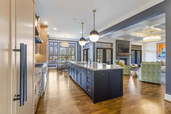 Navy blue kitchen with stained wood cabinets, large island and pendant lights looking into modern living room