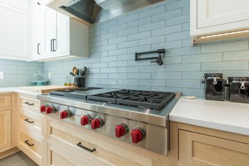 Wolf range with red knobs in a blue subway tile kitchen with white oak cabinets and white countertops