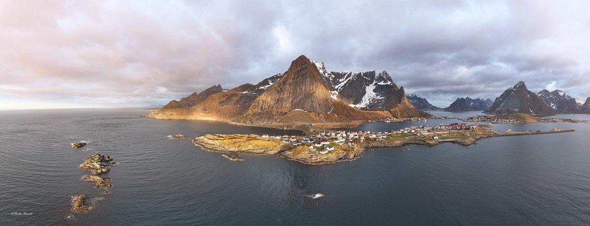 Aerial panorama of Reine and Sakrisøy villages beneath Olstinden peak in Lofoten, with fjords and islands stretching into the Norwegian Sea