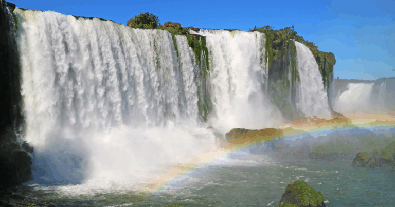 Vista panorâmica das Cataratas do Iguaçu, com quedas d’água imponentes cercadas por vegetação e um arco-íris formado pela névoa sobre o rio