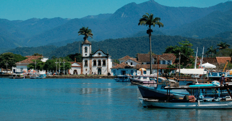 Vista de Paraty, no litoral do Rio de Janeiro, com igrejas históricas, casas coloniais e barcos coloridos ancorados no cais, tendo a serra ao fundo