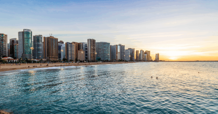 Vista da Praia de Meireles em Fortaleza ao entardecer, com o mar calmo refletindo o céu dourado e prédios modernos à beira-mar.