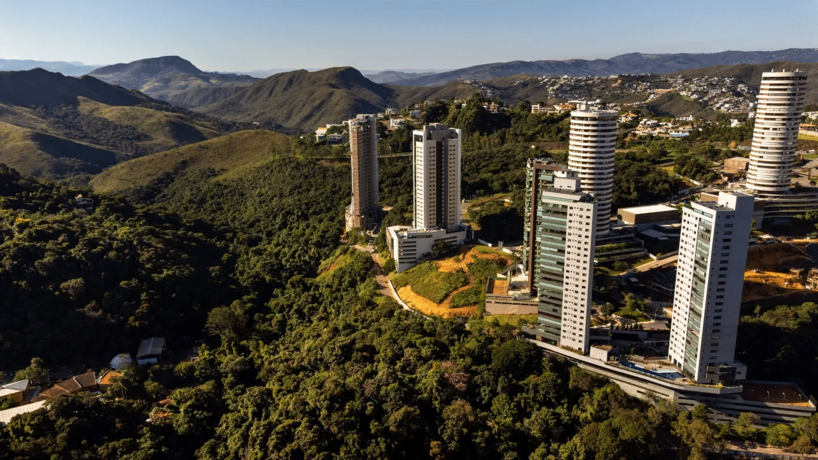 Vista aérea do bairro Belvedere, em Nova Lima (MG), mostrando prédios modernos em meio à vegetação da Serra da Moeda e montanhas ao fundo, sob céu limpo e ensolarado.