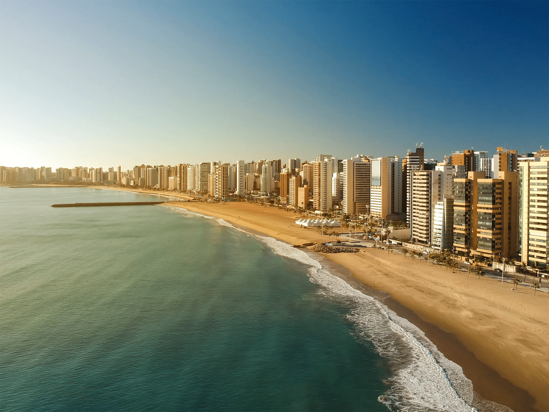 Vista panorâmica da Praia do Futuro, em Fortaleza (CE), com mar calmo, areia dourada e os modernos prédios da orla iluminados pelo sol