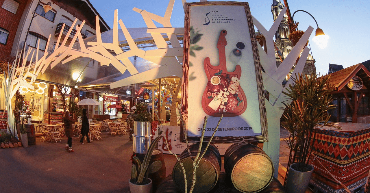 Festival de Cultura e Gastronomia de Gramado à noite, com mesas ao ar livre, decoração iluminada e cartaz do evento em destaque na rua central da cidade