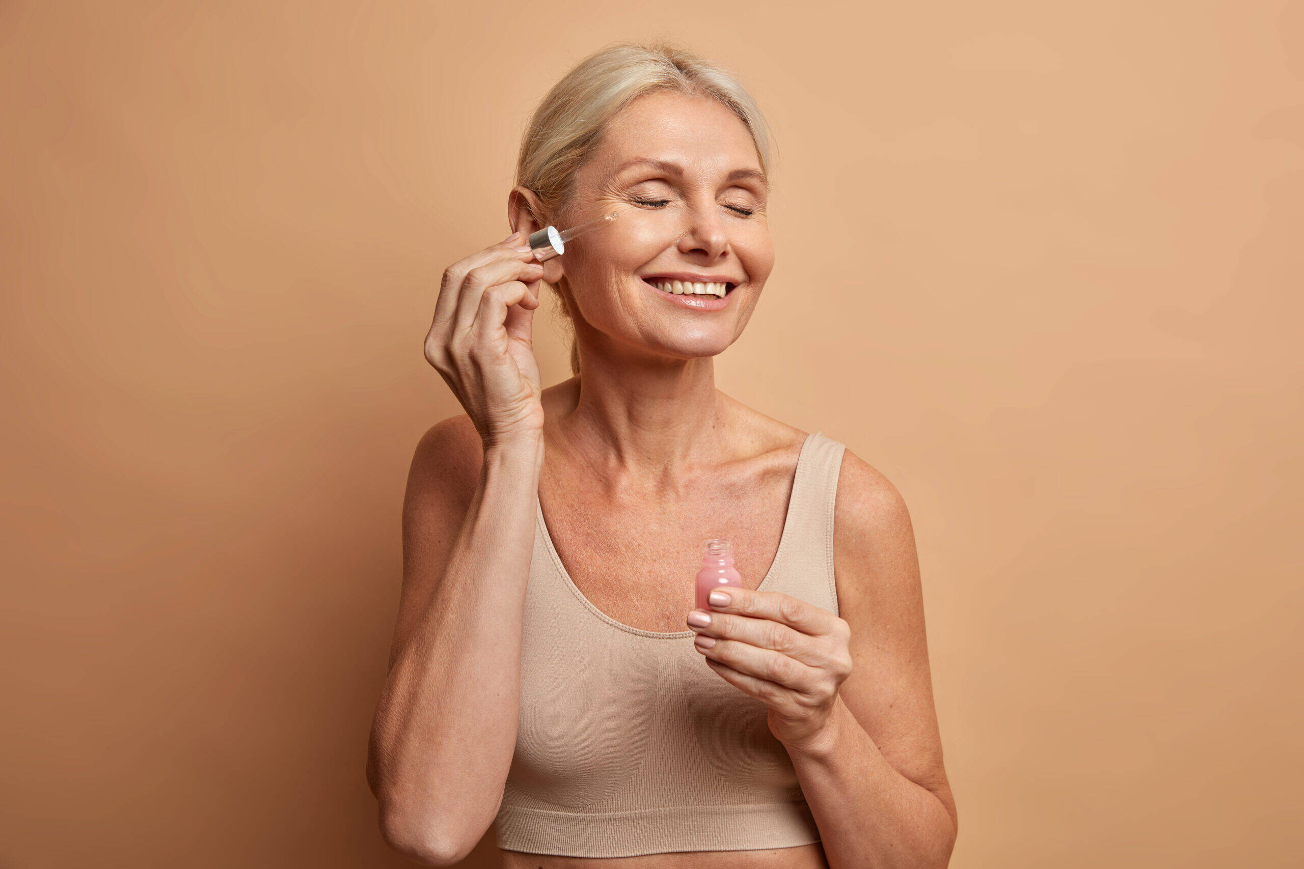 Pretty satisfied mature woman puts cosmetic serum on her face has shiny healthy skin closes eyes with satisfaction holds pipette of lactic acid wears top isolated over beige background. Beauty therapy