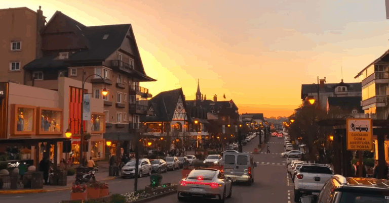 Rua Coberta e centro de Gramado ao entardecer, com luzes acesas, carros e pedestres em meio à arquitetura europeia sob um pôr do sol dourado