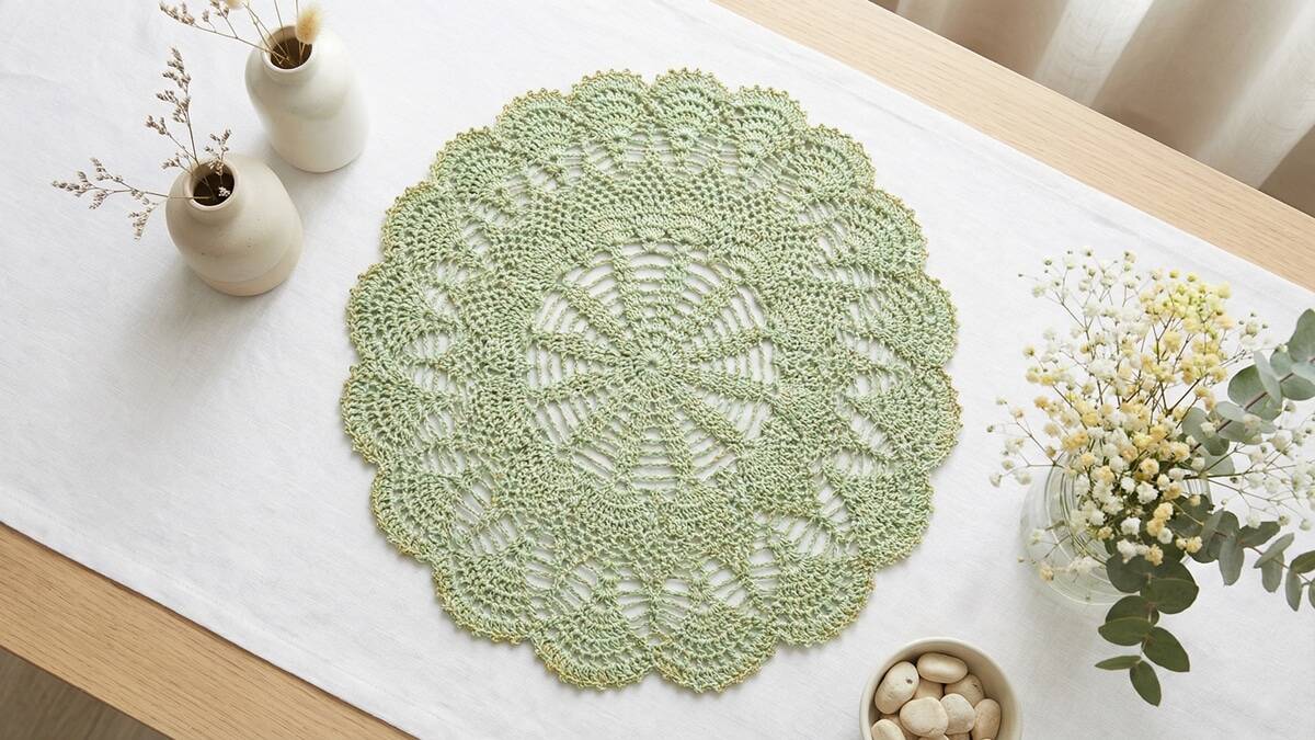 Mesa com centro de crochê redondo verde claro e detalhes dourados sobre toalha branca em composição elegante