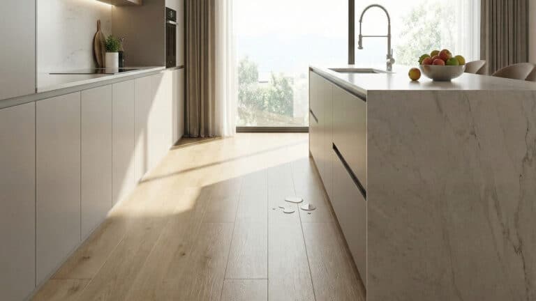 cozinha moderna e luxuosa, iluminada por luz natural vinda de uma grande janela. O destaque é o piso SPC de padrão madeira clara, que apresenta pequenas poças de água acumuladas na superfície próximo à ilha central de mármore branco, demonstrando sua impermeabilidade. Armários cinza minimalistas e uma bancada com pia e torneira completam o cenário.