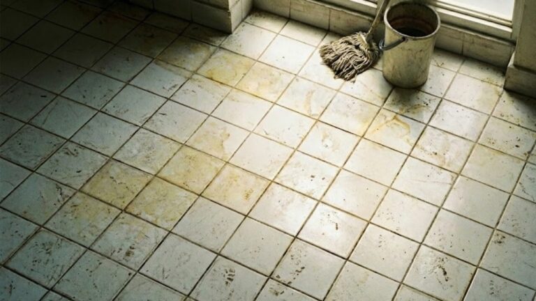 Piso de azulejos brancos encardido com manchas amareladas e marcas de pegadas, mostrando um balde e um esfregão de limpeza encostados na parede ao fundo.