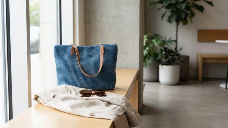 Bolsa de croche artesanal azul sobre banco de madeira clara com blazer neutro e oculos de sol em ambiente iluminado naturalmente