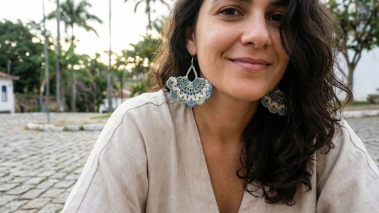 Mulher sorrindo usando brincos artesanais de croche em tons coloridos com trama rendada em ponto leque, ambiente externo com luz natural