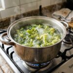 Panela de aço inox sobre a chama acesa de um fogão a gás, com flores de brócolis submersas em água fervente borbulhante, emitindo vapor espesso.