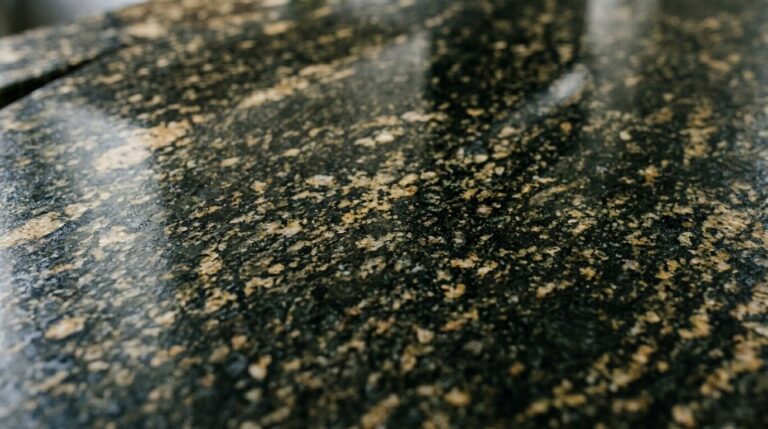 Fotografia em macro (close-up extremo) revelando os detalhes da textura de uma bancada de granito escuro. A superfície polida reflete a luz, destacando seus veios naturais e pequenos pigmentos minerais em tons de dourado, verde e preto.
