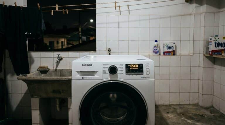 Máquina de lavar roupas branca de abertura frontal instalada em uma área de serviço azulejada, ao lado de um tanque de cimento, com produtos de limpeza ao fundo em ambiente noturno.