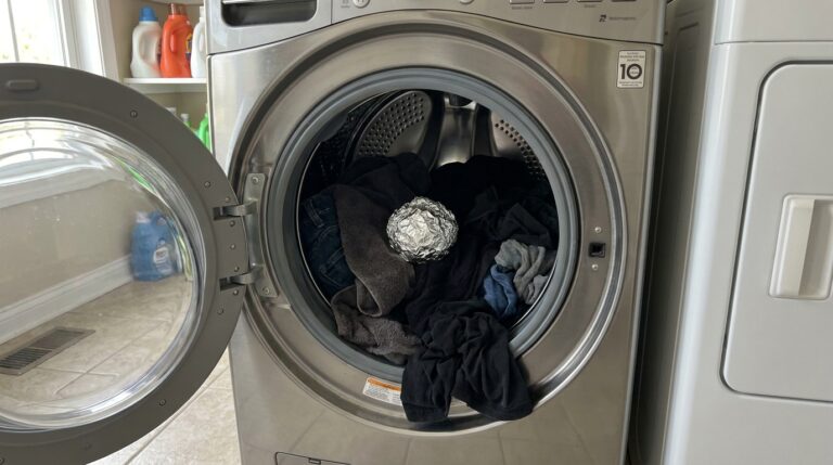 Tambor de aço inox de uma máquina de lavar roupas com a porta frontal aberta. O interior está preenchido com peças de roupas escuras e há uma bola de papel alumínio amassada centralizada sobre elas.
