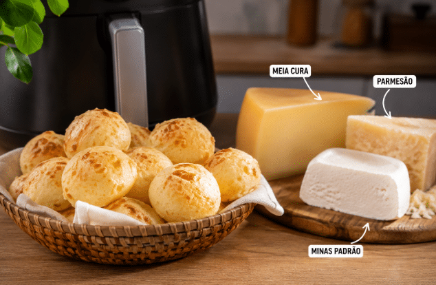 Cesta com pães de queijo dourados sobre mesa de madeira ao lado de uma tábua com três queijos — meia cura, parmesão e minas padrão — em ambiente de cozinha com Air Fryer ao fundo.