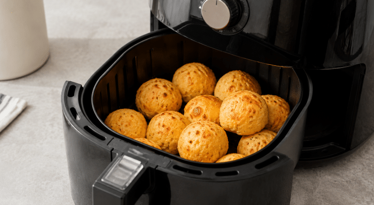 Pães de queijo dourados dentro da cesta de uma Air Fryer preta aberta sobre bancada clara de cozinha.