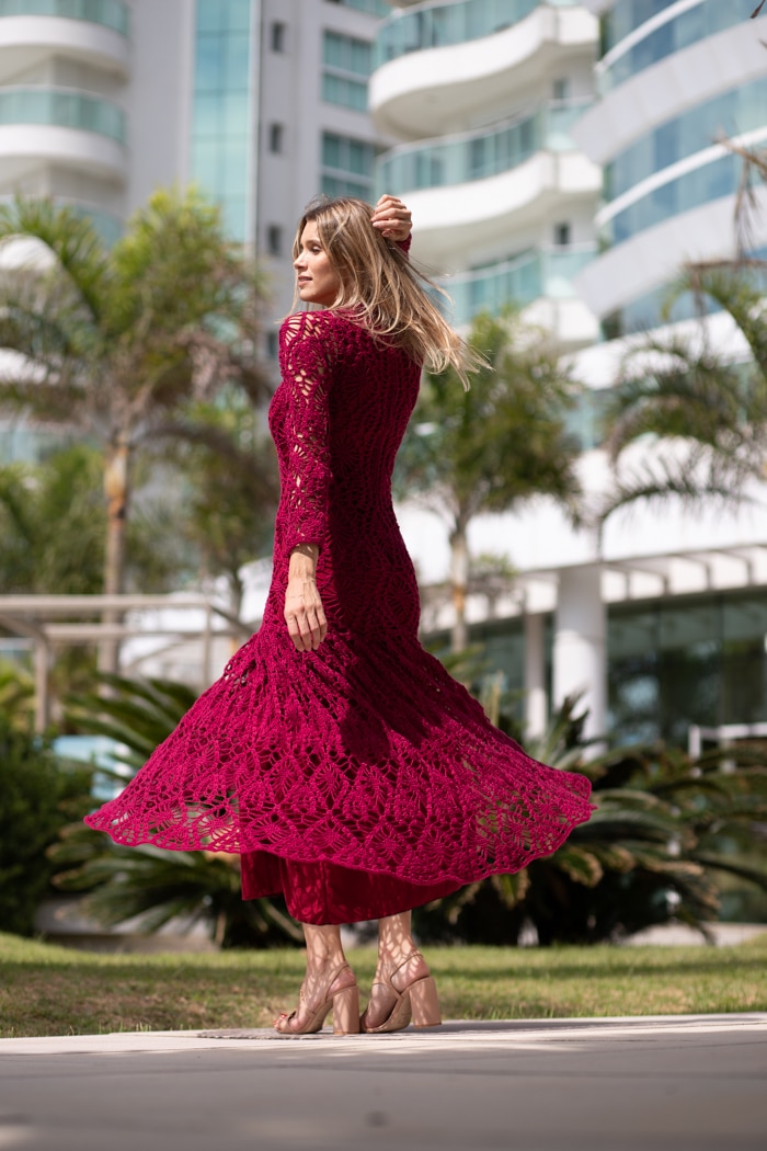 Manequim usando vestido longo de crochê bordô com mangas longas, gola alta e saia ampla em ambiente claro e sofisticado.
