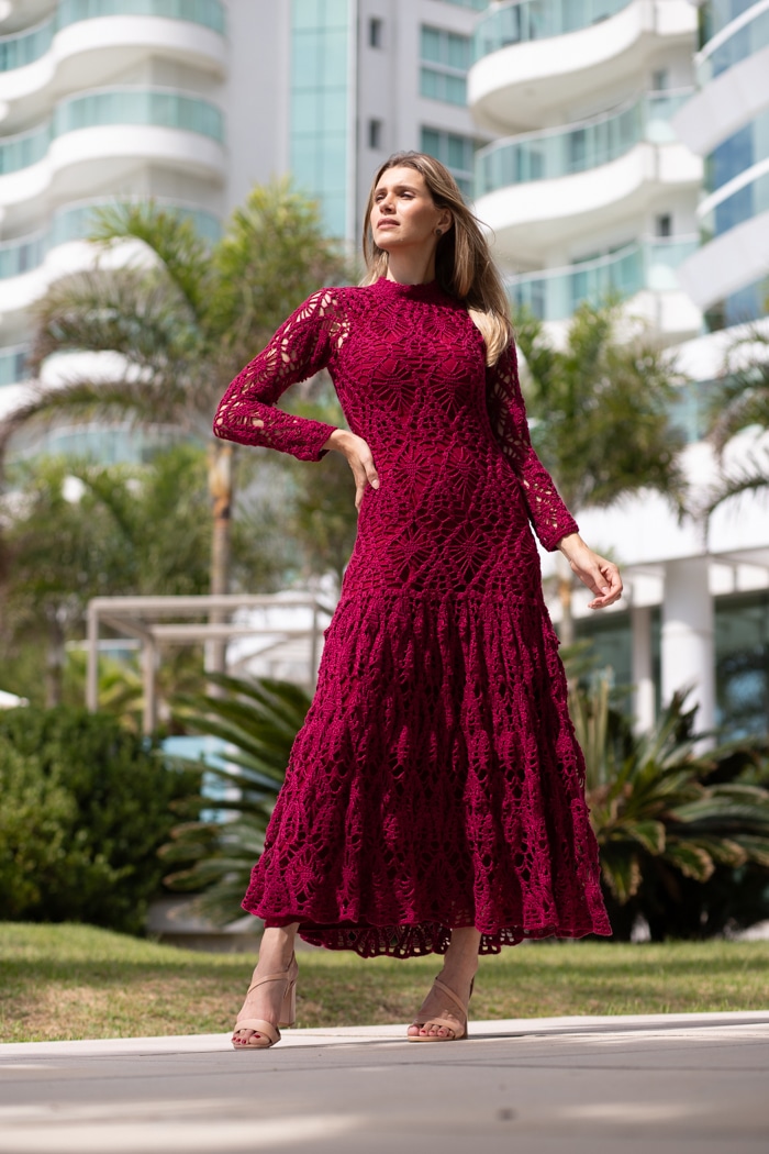 Manequim usando vestido longo de crochê bordô com mangas longas, gola alta e saia ampla em ambiente claro e sofisticado.
