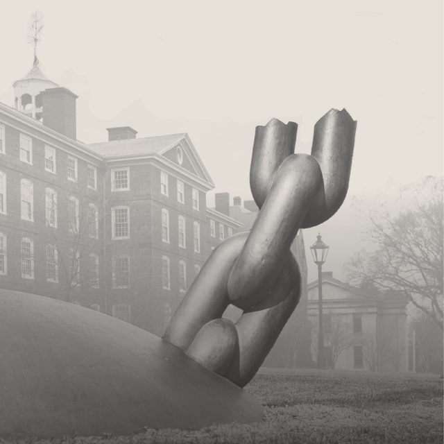 A grayscale image of a sculpture of a ball and broken chain protruding from grass. Trees and buildings are in the background. 