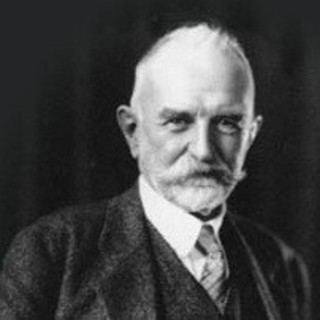 A grayscale photo portrait of George Herbert Mead. He has a neatly manicured beard with a moustache that sticks out to the sides. He is wearing a tweed suit with a tie. He is smirking and looking directly at the camera. Photo Credits: date and photographer unknown