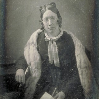 A faded and slightly cracked photo portrait of Catherine Beecher. She has a light-colored scarf across her shoulders, a dark dress, and a light-colored smaller neck-scarf. She is holding a piece of paper in her lap in one hand, and the other arm is rested on a table while holding a pen. The front/top of her hair has two neat space buns on either side of her head and the rest of her hair coils along her cheeks in a sort of short bob. She is looking directly at the camera with a very bored or exhausted facial expression. Photo Credit: possibly by W & F Langenheim (1848). Source: Wikimedia Commons.