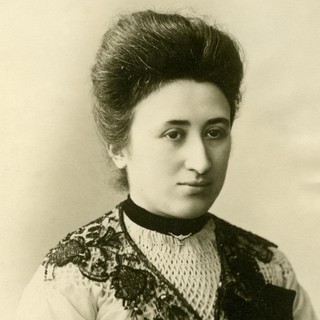 A sepia-toned photograph portrait of Rosa Luxemburg. She is wearing a high-neck dress with a v-neck-shaped lace detail as an accent. Her hair is neatly and voluminously coifed into an up-do. She is looking slightly to the right of the camera and she looks a bit bored or disinterested. Photographer: Karl Pinkau. Year: 1900. Source: Wikimedia Commons