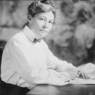 A gray-scale photograph of Frances Kellor sitting at a table with an open book. Her hands are rested on top of the book while she looks over her right shoulder at the photographer. She is wearing a light-colored collared shirt with a bow-tie. Her hair is pulled up in a loose, casual bun. Photo Credits: Bain News Service, 1910.