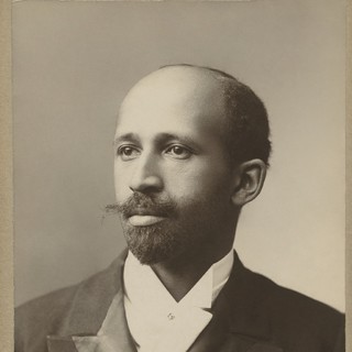 A sepia toned photo of W.E.B. Du Bois. He is a light-skinned Black man with a balding head, a moustache that protrudes sideways, and a narrow beard. 