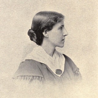 a sepia-toned photographed portrait of Charlotte Perkins Gilman. She is wearing a dress with puffy shoulder fabric and a white collar. Her hair is dark and pulled back in a low bun. She is turned all the way to the right, so you only see her side profile. Photo Credit: Barry and Marble, San Francisco, 1895 from commons.wikimedia.org